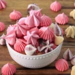 A small white bowl is filled with an assortment of colorful meringue kisses, predominantly in shades of pink, purple, and white. More meringues are scattered around the wooden surface, adding to the festive and sweet presentation.