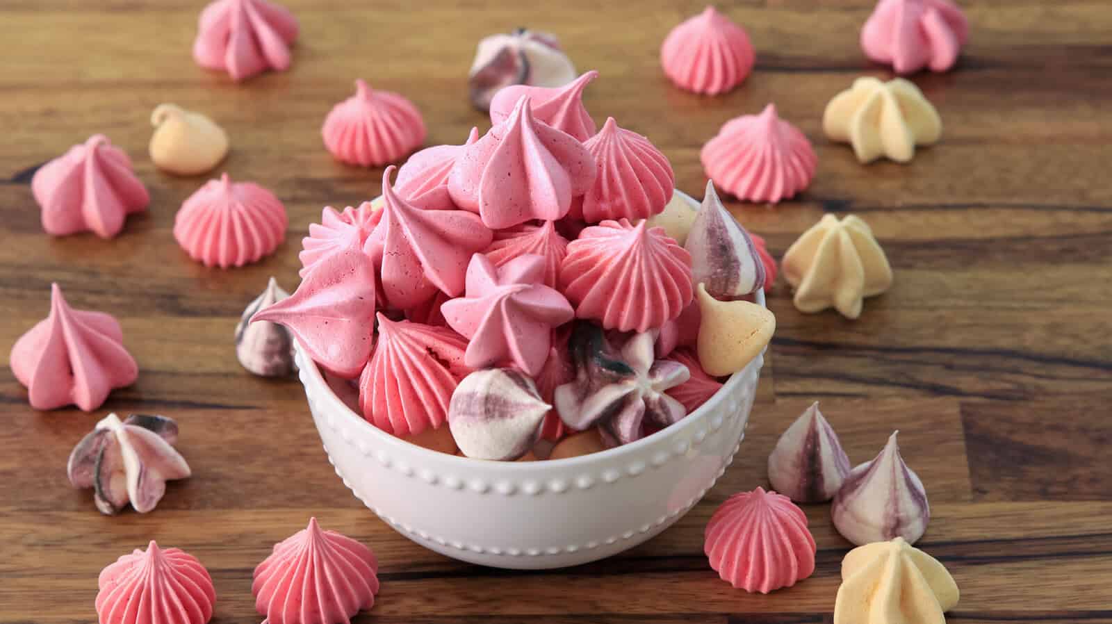 A small white bowl is filled with an assortment of colorful meringue kisses, predominantly in shades of pink, purple, and white. More meringues are scattered around the wooden surface, adding to the festive and sweet presentation.