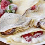 A plate of filled crepes dusted with powdered sugar. The crepes are folded and contain various fillings, including fresh strawberries, whipped cream, and possibly chocolate or banana slices. Some whole strawberries are also placed on the plate for garnish.