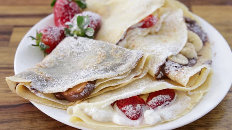 A plate of filled crepes dusted with powdered sugar. The crepes are folded and contain various fillings, including fresh strawberries, whipped cream, and possibly chocolate or banana slices. Some whole strawberries are also placed on the plate for garnish.