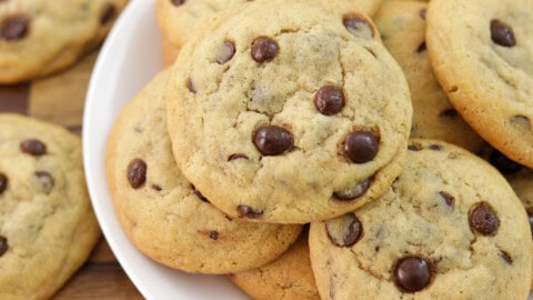 A close-up of a plate of chocolate chip cookies piled on top of each other. The cookies appear golden-brown and have visible chocolate chips. The plate is against a wooden background.