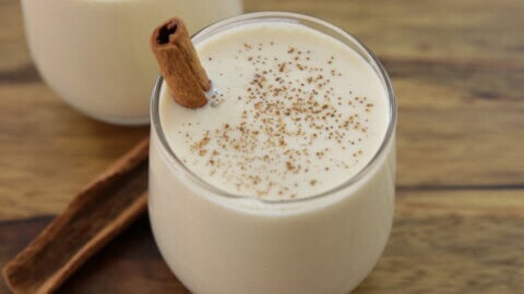 Two glasses filled with creamy eggnog are topped with ground nutmeg and a cinnamon stick. The drinks are set on a wooden surface with two additional cinnamon sticks lying beside the glasses, evoking a cozy holiday feel.