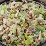 A close-up of a bowl filled with a hearty chicken salad. It contains diced grilled chicken, chopped lettuce, red onions, celery, raisins, and is dressed with a creamy sauce. The vibrant colors and diverse textures of the ingredients are clearly visible.