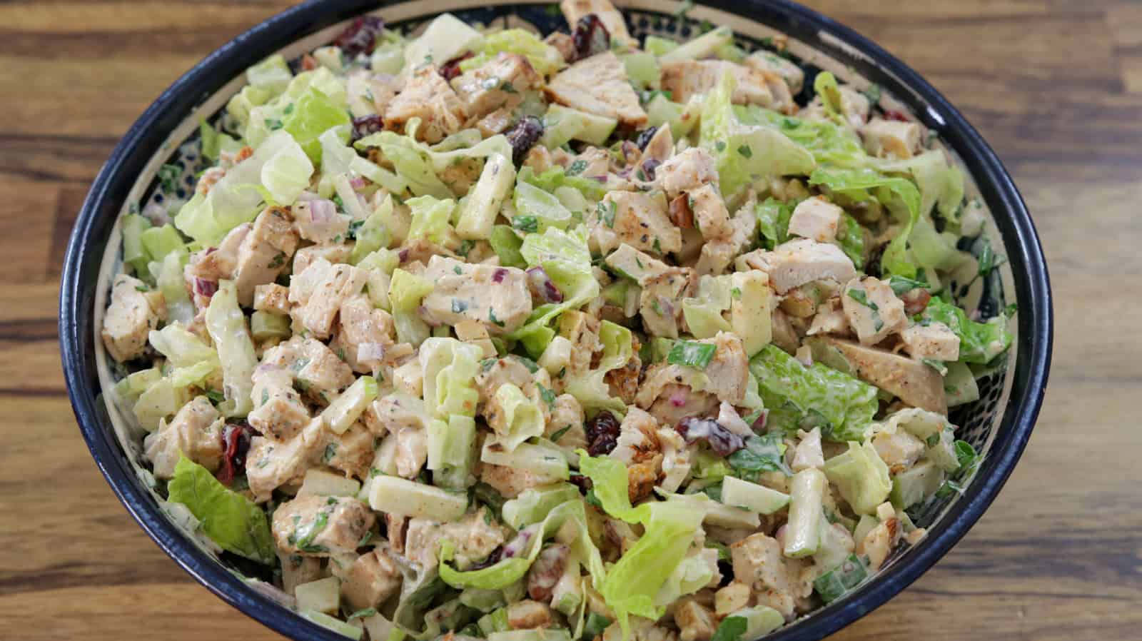 A close-up of a bowl filled with a hearty chicken salad. It contains diced grilled chicken, chopped lettuce, red onions, celery, raisins, and is dressed with a creamy sauce. The vibrant colors and diverse textures of the ingredients are clearly visible.