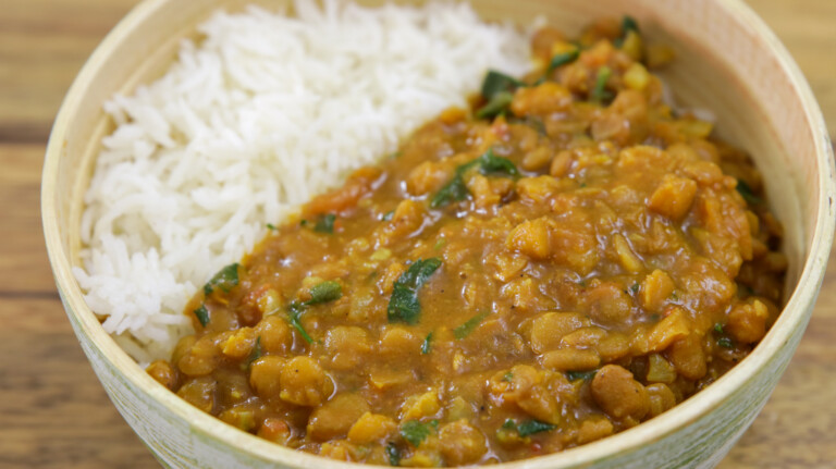 A bowl filled with white rice and a serving of lentil curry. The curry is yellow-orange in color and includes visible chunks of vegetables and green herbs. The bowl is placed on a wooden surface.