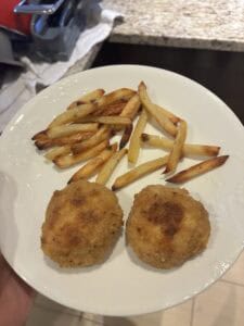 A white plate with two breaded, fried croquettes and a serving of golden, baked French fries, held over a kitchen counter.