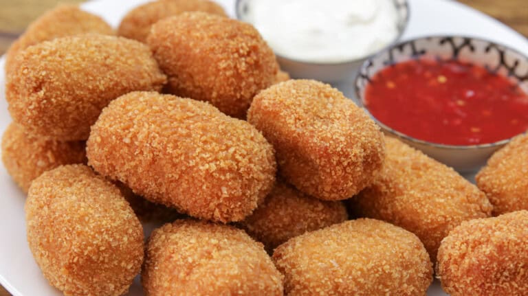 A plate of golden-brown, breaded chicken nuggets is arranged around two small bowls of dipping sauces: one with a white creamy sauce and the other with a red chili sauce. The plate is set on a wooden surface.