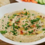 A white bowl filled with creamy baba ganoush, garnished with olive oil, chopped parsley, and paprika, sits on a wooden table. Pita bread, sliced cucumbers, and carrot sticks are visible in the background.