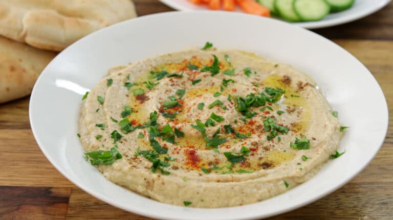 A white bowl filled with creamy baba ganoush, garnished with olive oil, chopped parsley, and paprika, sits on a wooden table. Pita bread, sliced cucumbers, and carrot sticks are visible in the background.