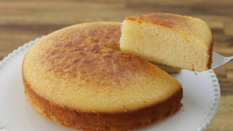 A hand holds a spatula lifting a slice of golden-brown cornbread from a round, evenly baked cornbread sitting on a white plate. The background features a wooden surface, slightly blurred.