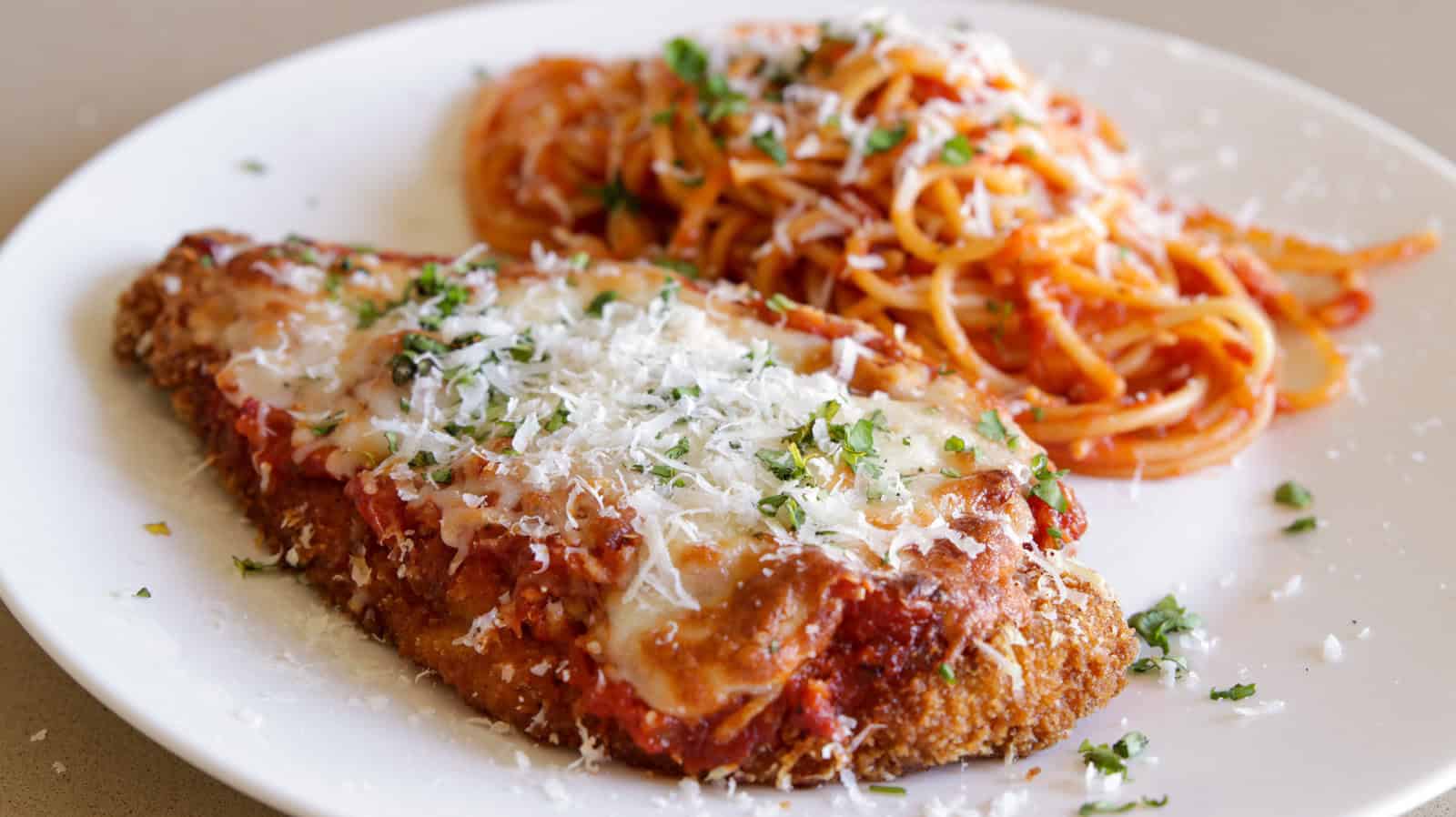 A plate with breaded chicken parmesan topped with melted cheese and marinara sauce, garnished with chopped parsley, next to a portion of spaghetti mixed with tomato sauce, sprinkled with grated cheese.