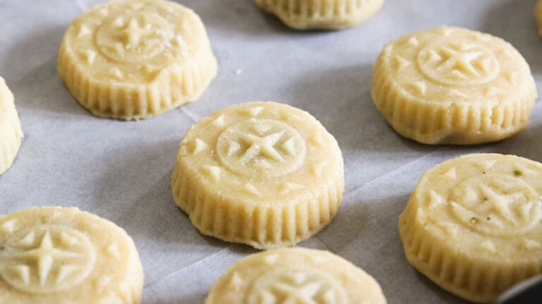 Left side: Several round, intricately patterned cookies on parchment paper. Right side: One of the same cookies on a plate, broken in half to reveal a filling of dates or nuts, with powdered sugar dusting on top. Maamoul recipe.