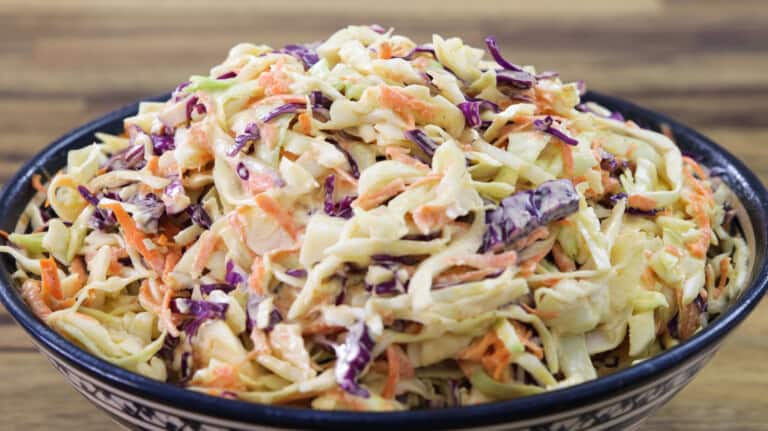 A close-up view of a bowl filled with coleslaw. The coleslaw is made up of shredded cabbage, both green and purple, as well as thin strips of carrots, all mixed together with a creamy dressing. The dish looks fresh and colorful.