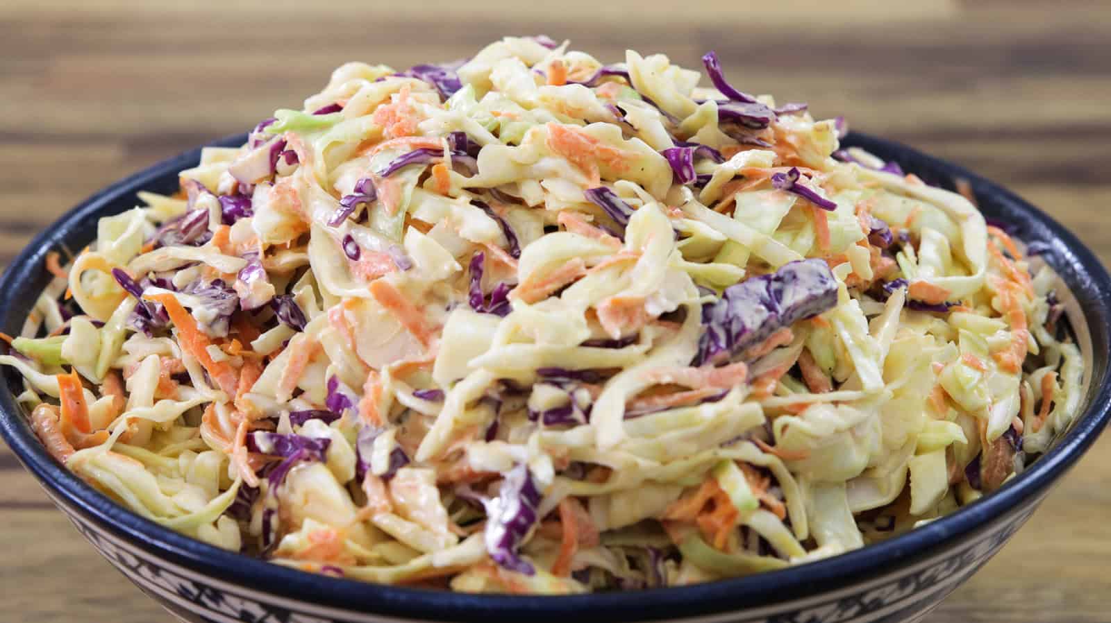 A close-up view of a bowl filled with coleslaw. The coleslaw is made up of shredded cabbage, both green and purple, as well as thin strips of carrots, all mixed together with a creamy dressing. The dish looks fresh and colorful.