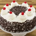 A round cake on a white pedestal stand, decorated with white frosting, chocolate shavings on the sides, a circle of chocolate shavings on top, and eight swirls of whipped cream each topped with a red cherry around the edge. The background is a wooden surface.