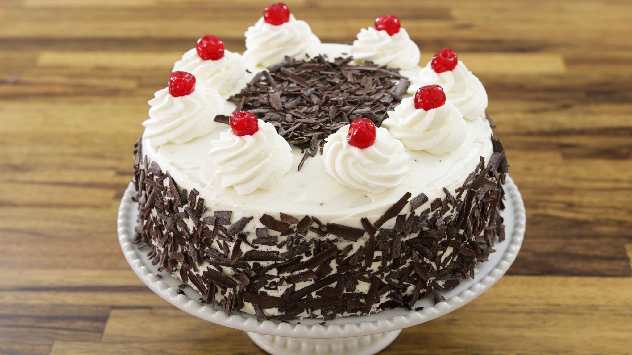 A round cake on a white pedestal stand, decorated with white frosting, chocolate shavings on the sides, a circle of chocolate shavings on top, and eight swirls of whipped cream each topped with a red cherry around the edge. The background is a wooden surface.