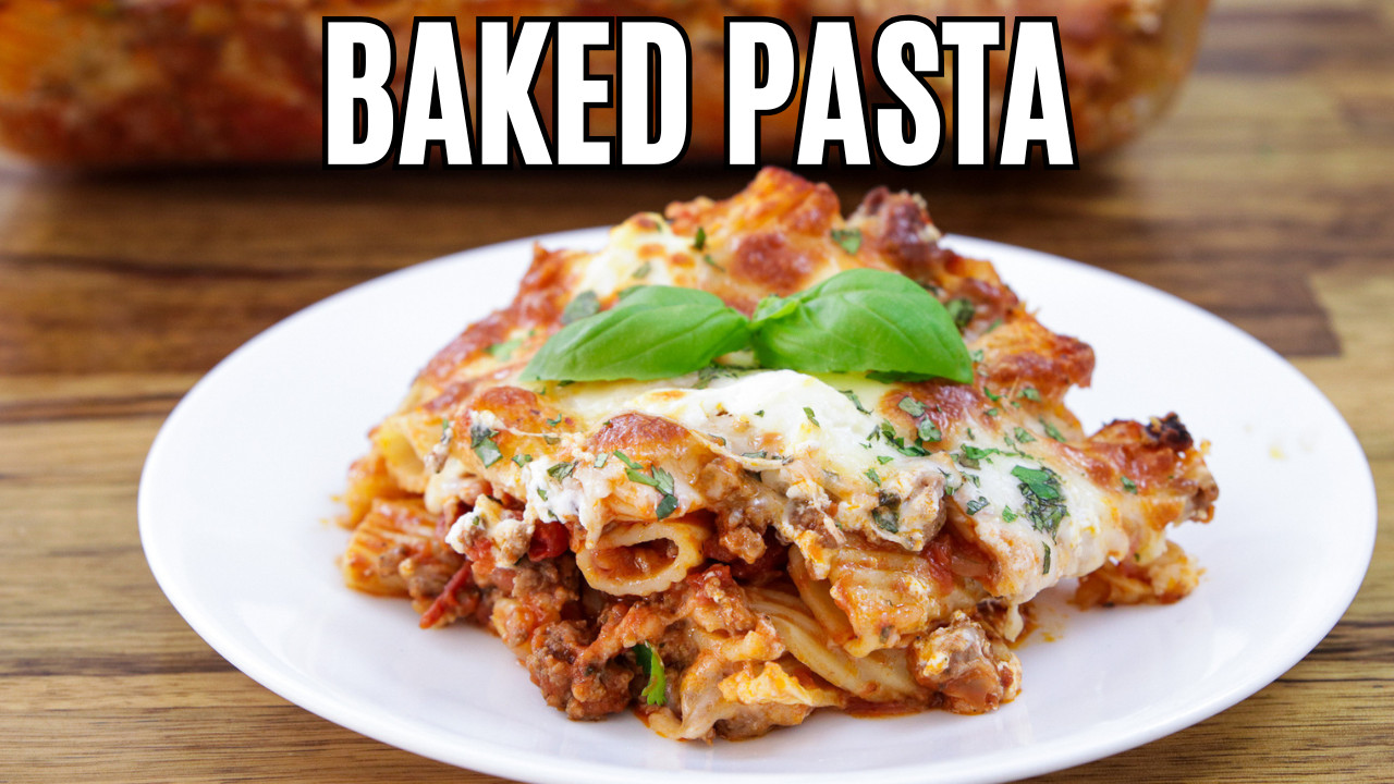 A serving of baked ziti with melted cheese and basil on top is placed on a white plate. In the background, a glass baking dish is filled with more baked ziti. The dish is set on a wooden table.