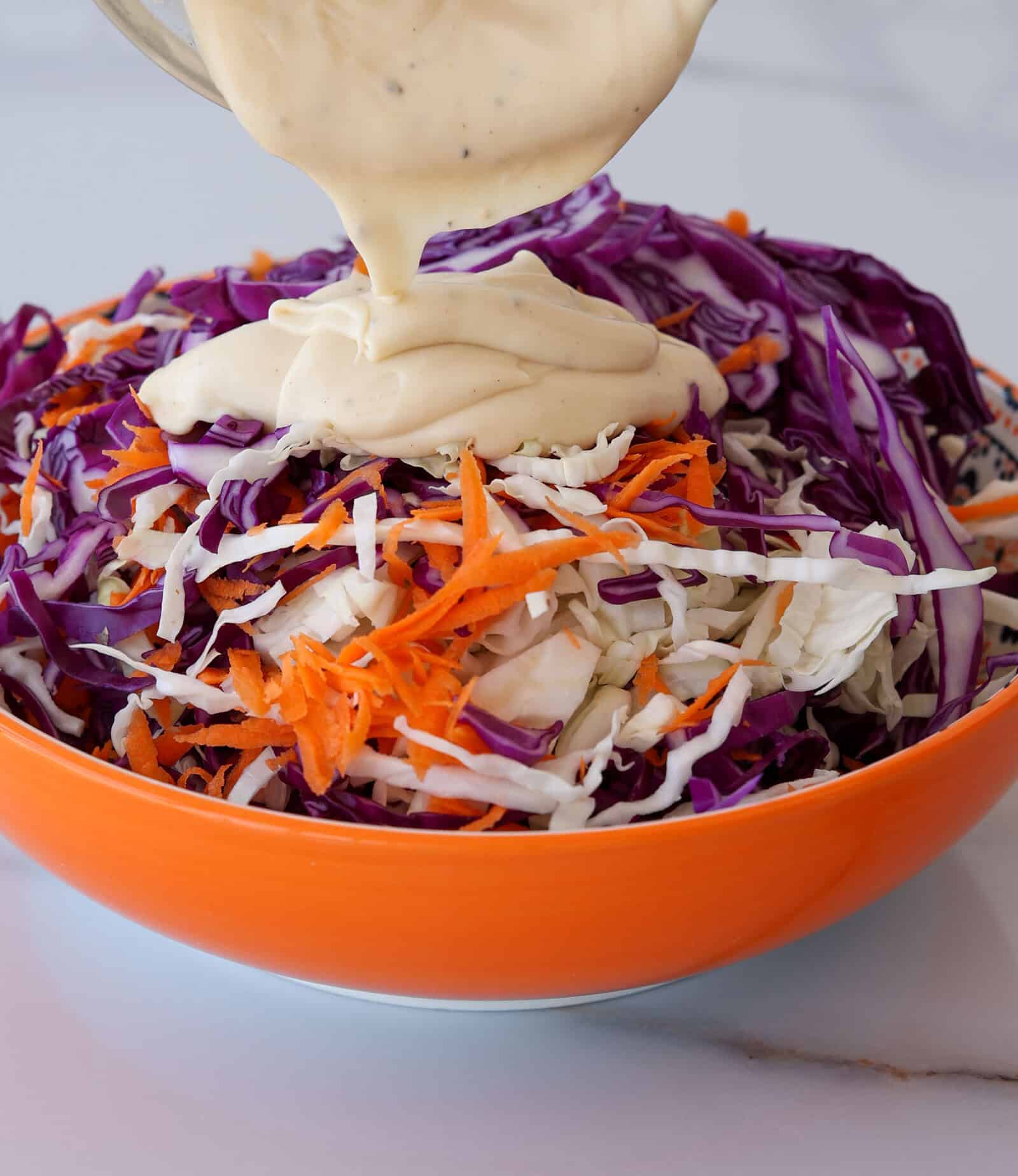Shredded purple cabbage, white cabbage, and carrots in an orange bowl with creamy dressing being poured on top.