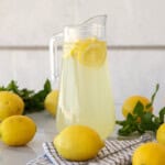 A clear glass pitcher filled with lemonade and lemon slices sits on a white surface, surrounded by whole lemons, green leaves, and a folded checkered cloth. The background is light and minimalistic.