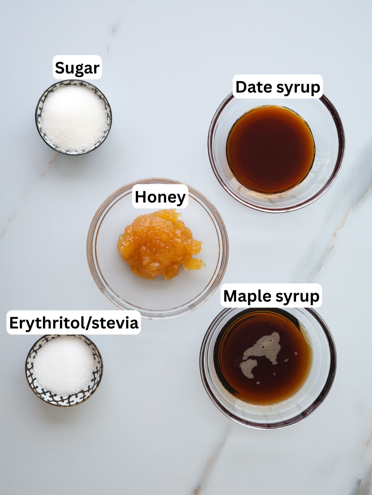 Five small bowls on a white surface, each labeled: sugar, date syrup, honey, maple syrup, and erythritol/stevia. The bowls contain different sweeteners in powdered, liquid, and paste forms.