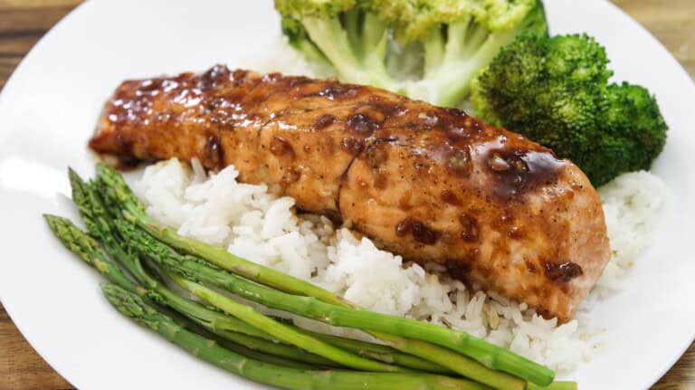 A plate of teriyaki-glazed salmon served on a bed of white rice, accompanied by steamed asparagus and broccoli florets. The dish is presented on a white plate, with the vegetables neatly arranged around the salmon.