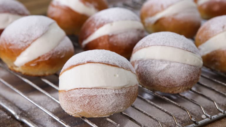 A batch of cream-filled doughnuts rests on a cooling rack. The doughnuts are covered with a light dusting of powdered sugar, and a generous amount of creamy filling is visible in the middle of each one.