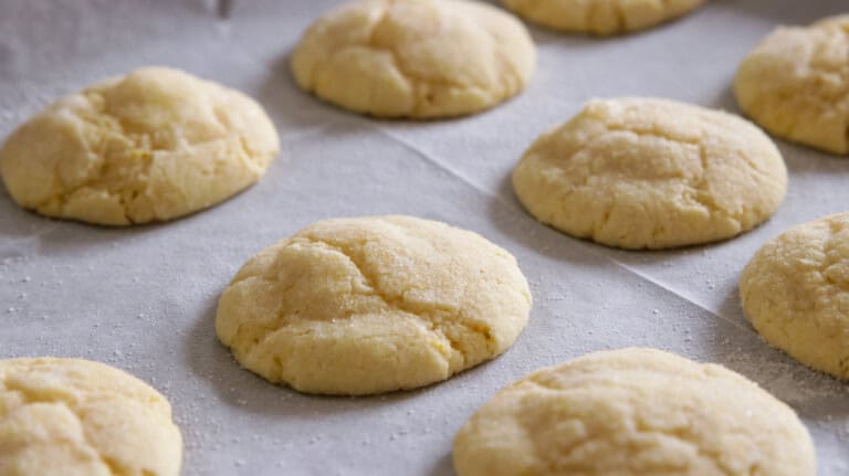 Freshly baked soft lemon cookies are arranged on a parchment-lined baking sheet. The cookies are round, slightly cracked on top, and lightly dusted with sugar.