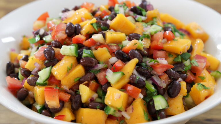 A white bowl filled with a colorful salad featuring mango chunks, black beans, diced red bell peppers, cucumbers, red onions, and chopped cilantro. The ingredients are mixed and displayed closely, highlighting the vibrant, fresh appearance of the dish.