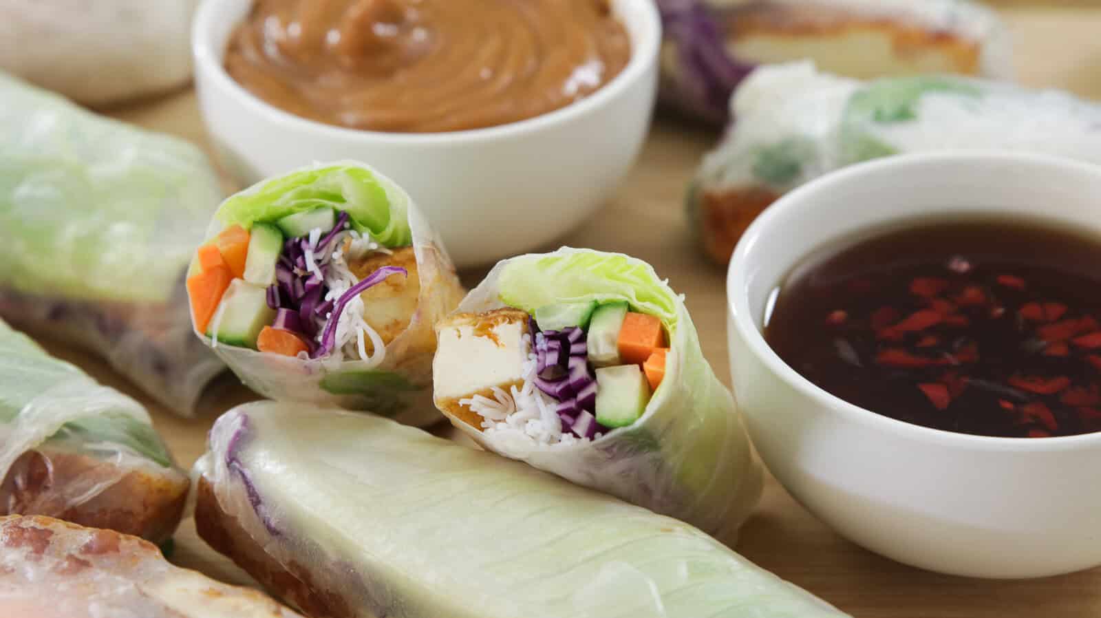 A wooden board displaying several fresh spring rolls, each filled with tofu, vegetables, and herbs wrapped in translucent rice paper. Two small bowls of dipping sauces, one with a brown peanut sauce and the other with a red chili sauce, are placed on the board.