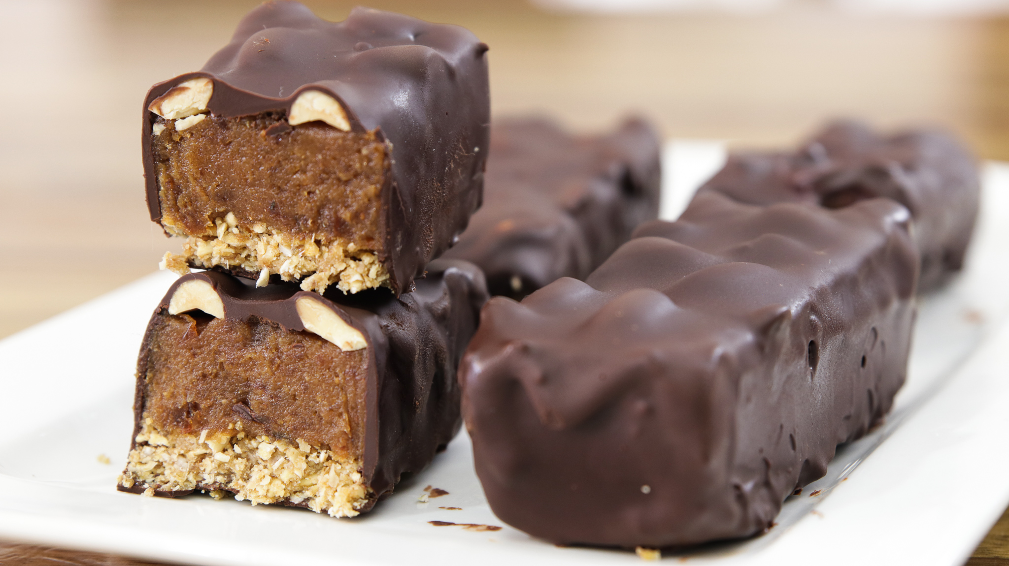 Close-up of chocolate-covered granola bars on a white plate, with one bar cut in half to show the interior, revealing nuts and a dense filling. The bars have a crunchy base and a smooth chocolate coating.