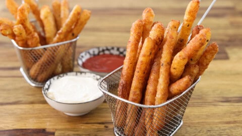 Two metal baskets filled with crunchy, golden-brown mashed potato sticks sit on a wooden table. One small bowl of marinara sauce and another small bowl of aioli dip are placed between the baskets.