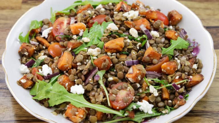 A colorful salad in a white bowl features a mix of lentils, arugula, cherry tomatoes, roasted sweet potatoes, red onions, and crumbled feta cheese. The ingredients are tossed together, creating a vibrant and healthy dish.