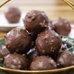 A decorative bowl filled with chocolate-coated truffles stacked in a pyramid shape. The truffles have a textured surface, suggesting a crunchy coating. In the background, more truffles can be seen on a plate. The setup is on a wooden table.