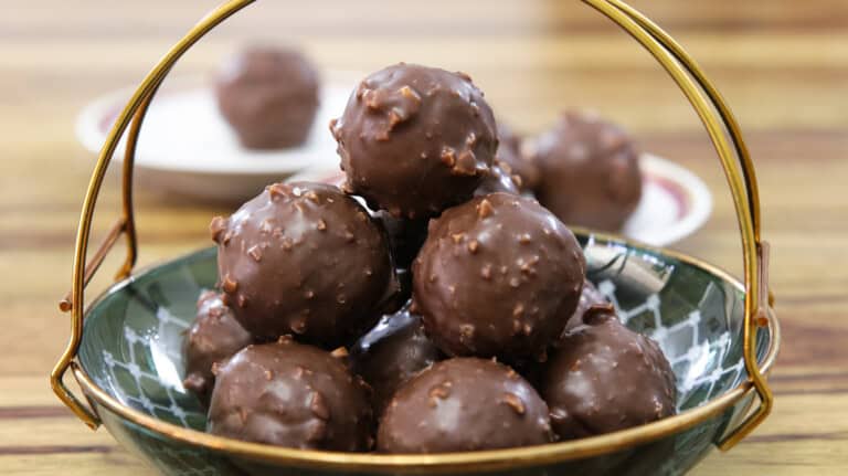 A decorative bowl filled with chocolate-coated truffles stacked in a pyramid shape. The truffles have a textured surface, suggesting a crunchy coating. In the background, more truffles can be seen on a plate. The setup is on a wooden table.