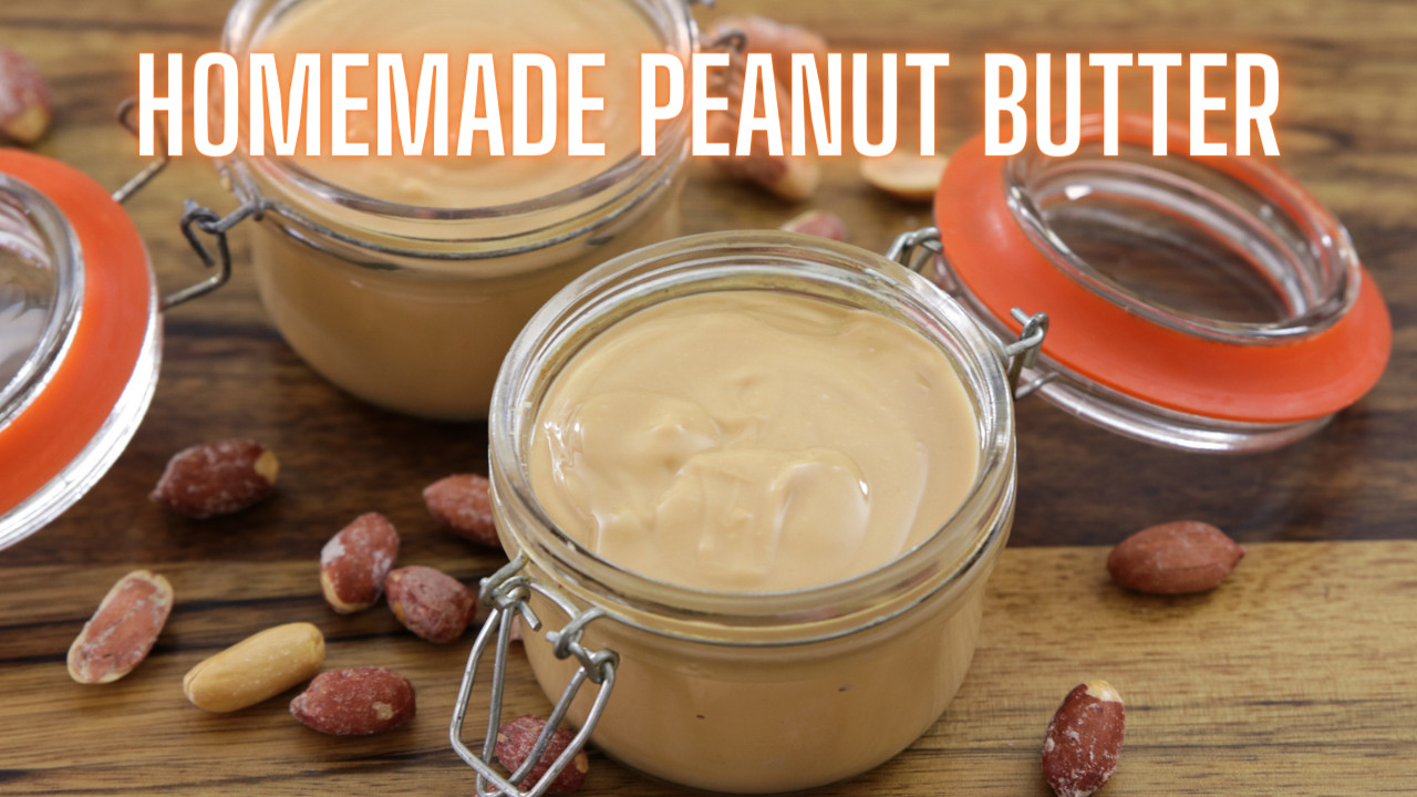 Three glass jars of creamy homemade peanut butter are displayed on a wooden surface. The jars have orange sealing rings, and some are open. Peanut shells and whole peanuts are scattered around the jars. The text "HOMEMADE PEANUT BUTTER" is written above.