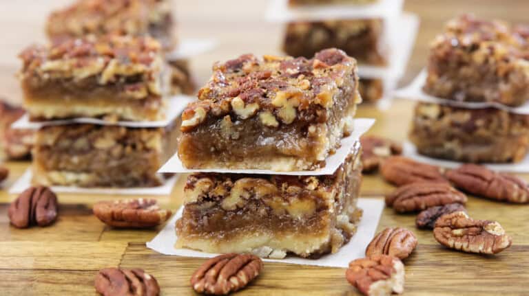 A close-up of two stacked pecan pie bars on a wooden surface. The bars feature a crumbly crust, a gooey caramel-like filling, and a generous topping of pecans. Additional pecan pie bars and loose pecan nuts are scattered in the background.