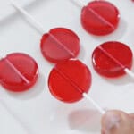 Seven red, round lollipops arranged in a grid pattern on a white rectangular plate. A hand is holding the stick of one lollipop at the bottom right of the image.