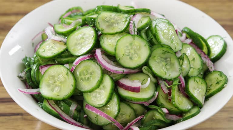 A white bowl filled with crunchy cucumber salad, red onions, and chopped fresh herbs, likely dill, on a wooden surface.