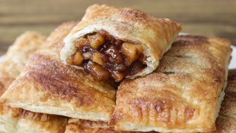 A close-up of a plate topped with four golden-brown rectangular pastries. The pastries have a flaky texture and are arranged in a neat stack. The plate is placed on a wooden surface, enhancing the warm, inviting appearance of the pastries.