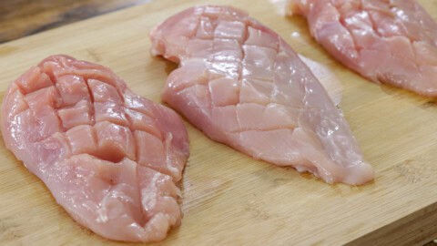 Three raw, scored chicken breasts are placed on a wooden cutting board. The scoring creates a grid-like pattern on the surface of the chicken pieces. The background is a rustic wooden table.