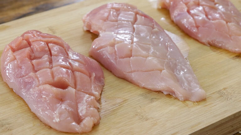 Three raw, scored chicken breasts are placed on a wooden cutting board. The scoring creates a grid-like pattern on the surface of the chicken pieces. The background is a rustic wooden table.