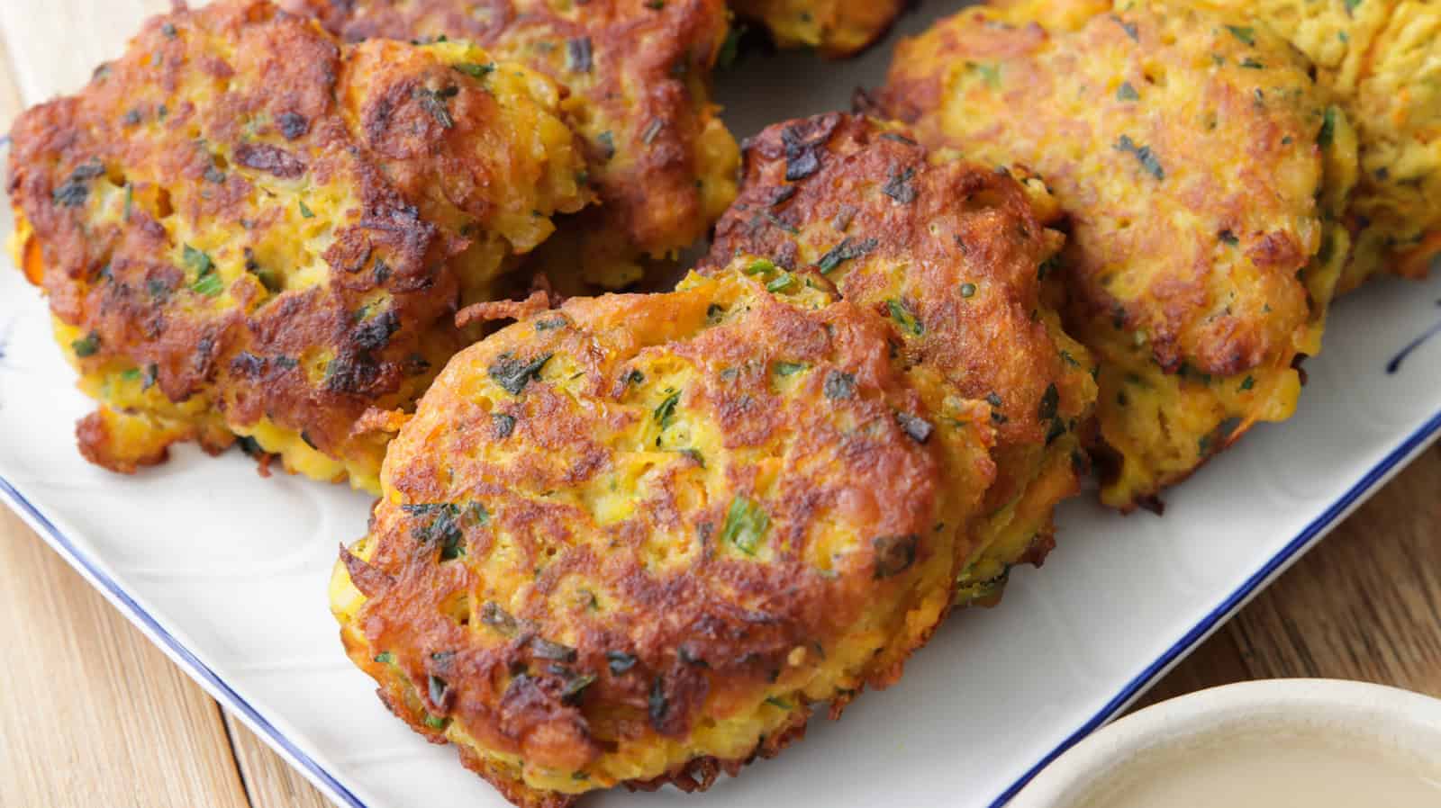 A close-up of a plate of golden-brown vegetable fritters. The fritters are slightly crispy on the outside, with visible flecks of green herbs and orange carrot pieces. The plate is set on a wooden surface with a small bowl of dipping sauce partially visible.