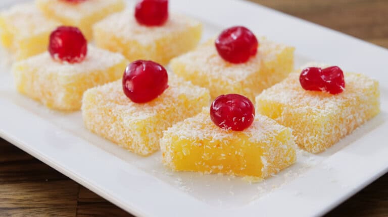 Squares of yellow cake topped with desiccated coconut and red glace cherries are arranged on a white rectangular plate. The yellow cakes have a smooth texture and are visually contrasted by the white coconut and bright red cherries.