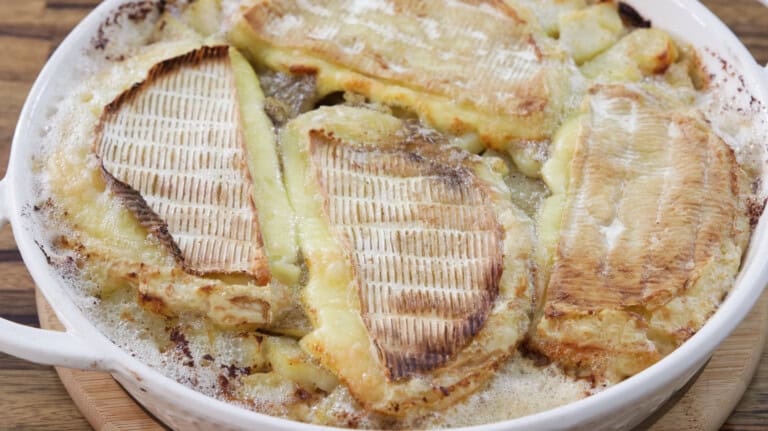 A round, white dish filled with a freshly baked gratin featuring golden, melted cheese slices on top of thinly sliced potatoes. The dish has browned edges, suggesting it has been oven-baked to a crispy finish. The background is a wooden table.