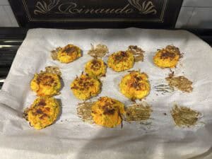 Ten golden-brown vegetable patties rest on a sheet of parchment paper atop a baking tray, with some browned spots indicating they've been oven-baked. The oven brand "Rinaudo" is visible in the background.