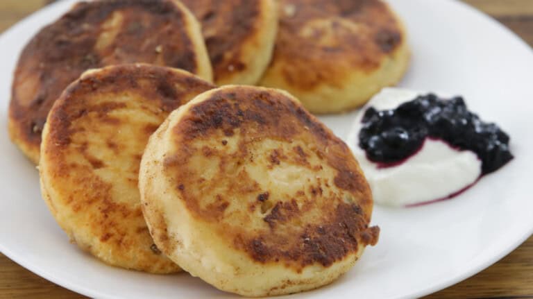 Five golden-brown syrniki pancakes are served on a white plate with a side of white cream topped with a spoonful of dark berry sauce.