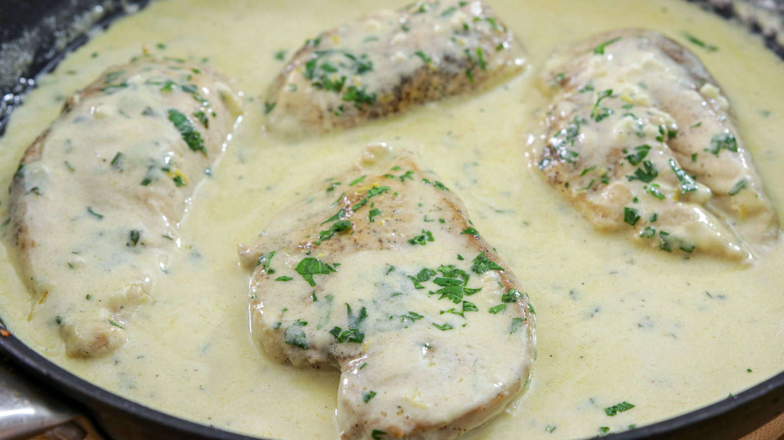 Chicken breasts cooking in a creamy sauce with herbs in a skillet. The sauce looks rich and smooth, generously coating the chicken, which is garnished with chopped parsley.