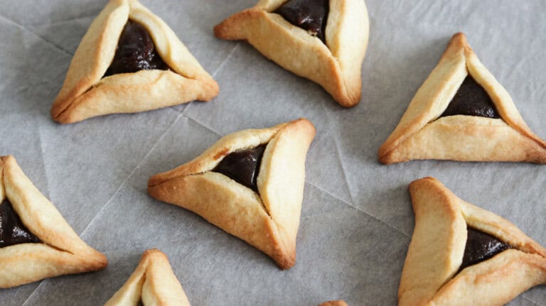 A close-up of several Hamantaschen cookies on a baking sheet. These triangular pastries are filled with a dark jam and dusted with powdered sugar. The cookies are arranged neatly in rows.