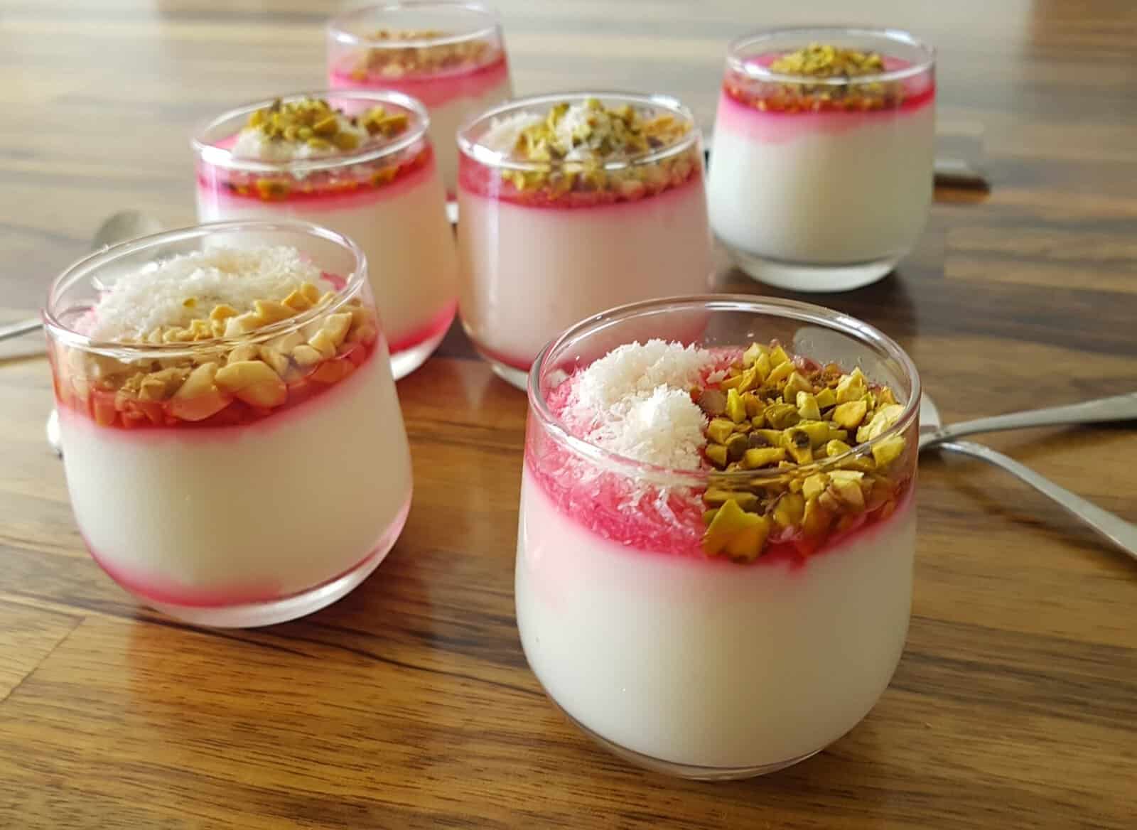 six cups of a malabi pudding dessert topped with a red syrup and chopped pistachios and coconut