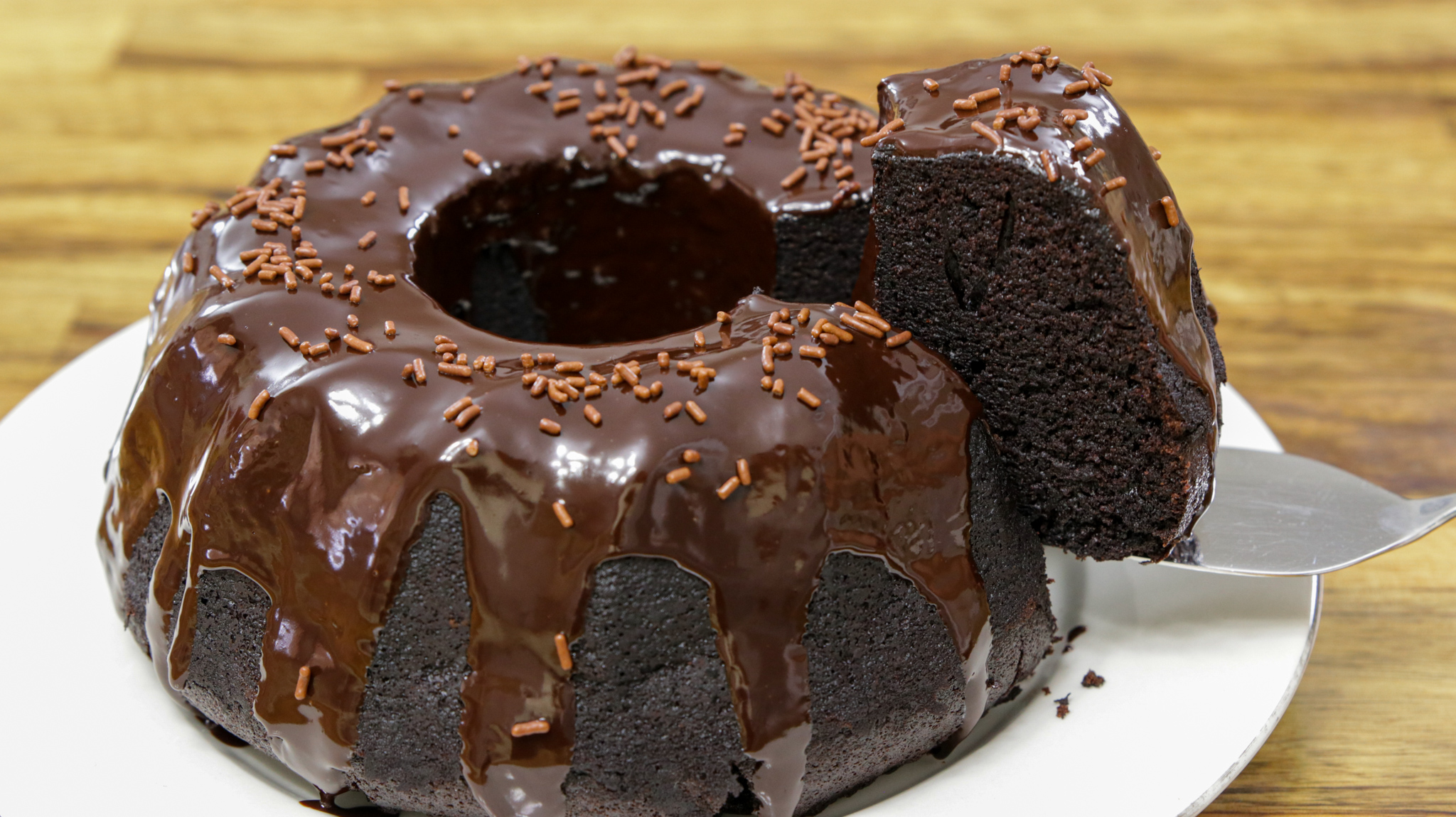 A rich chocolate bundt cake topped with glossy chocolate ganache and chocolate sprinkles sits on a white plate. A slice is being lifted out with a silver cake server, revealing the moist, dark interior. The background is a wooden table.